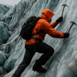A determined ice climber in an orange jacket and black backpack ascends a steep, jagged glacier wall using ice axes, surrounded by dramatic blue-toned ice formations under a cloudy sky.