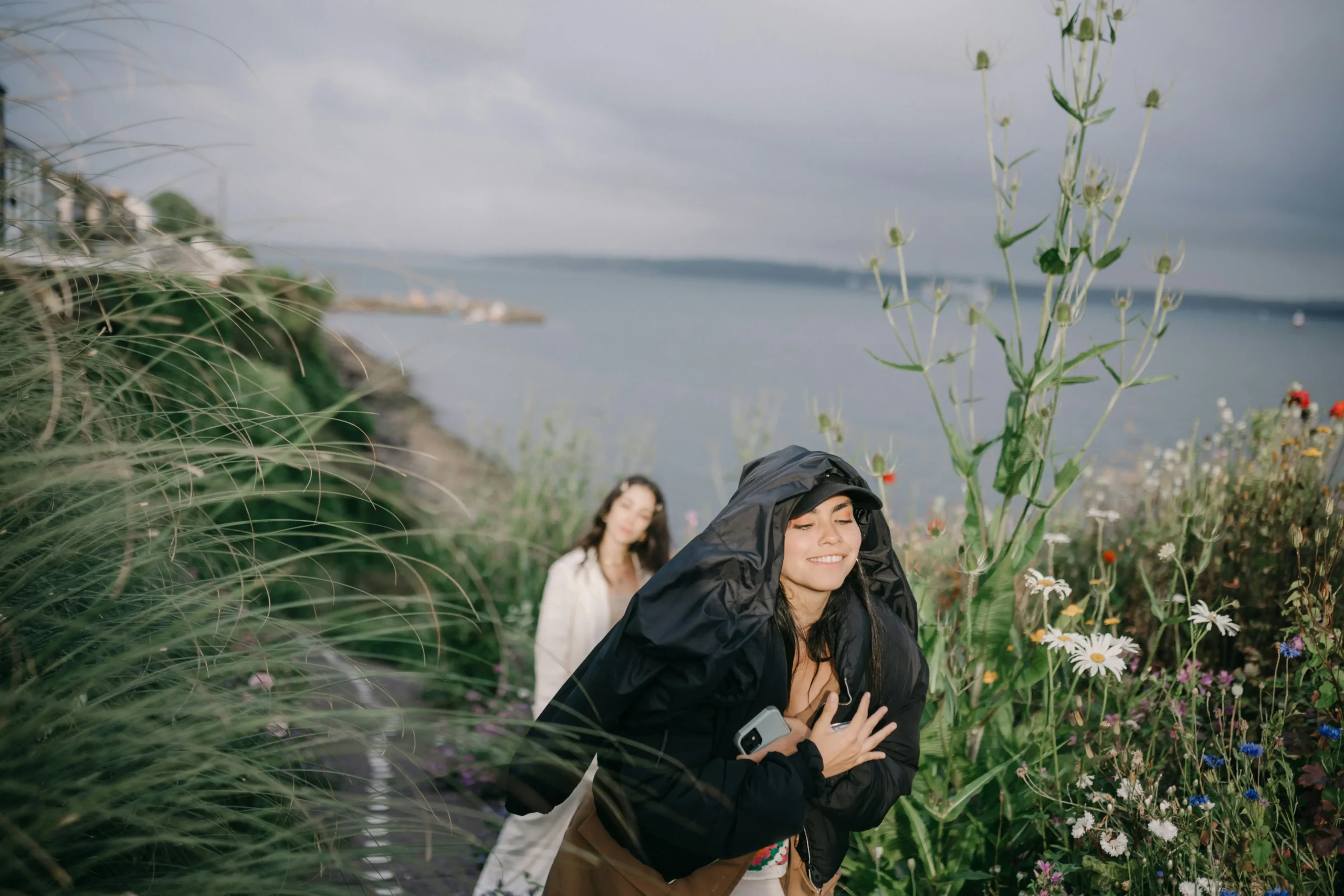 Two women walking along a scenic coastal path, one smiling with a jacket over her head, symbolizing a positive travel mindset despite the weather.