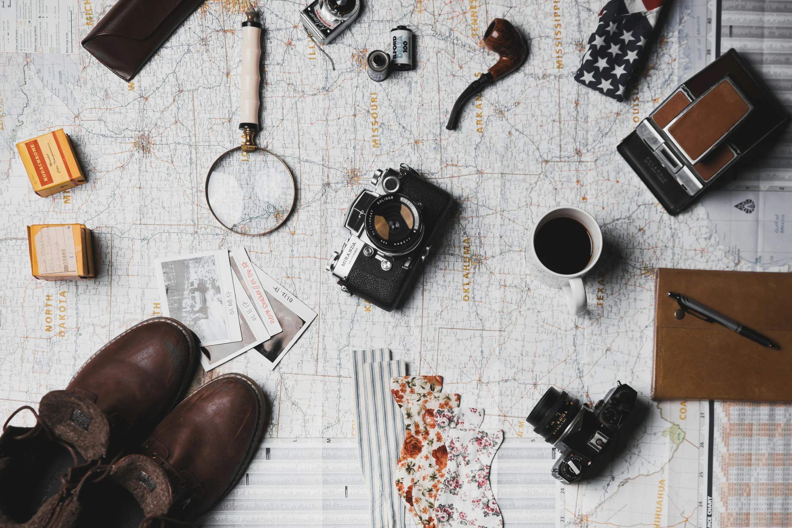 Overhead view of vintage travel items including cameras, maps, boots, passport, and journal arranged on weathered map background
