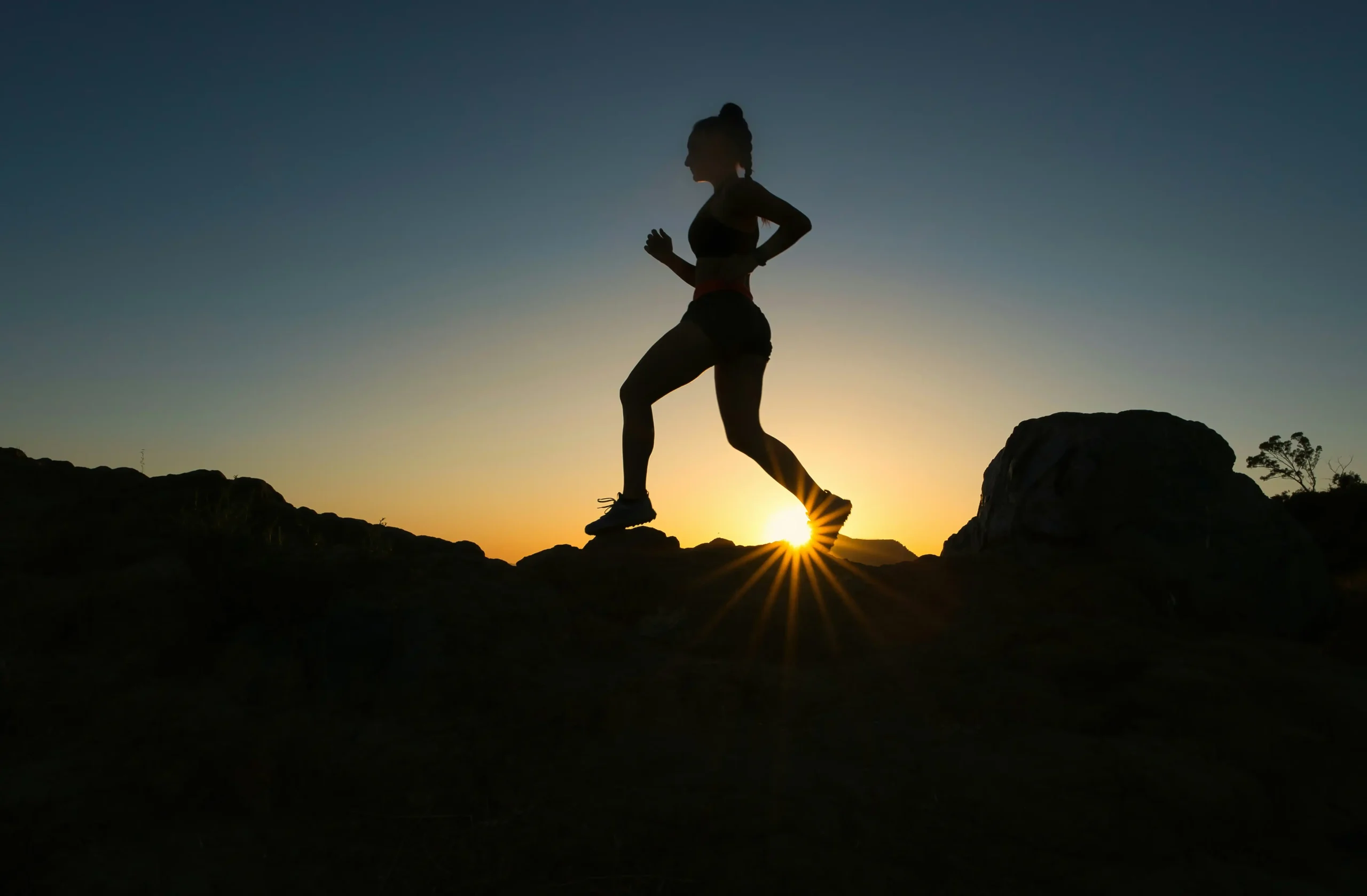 Silhouetted female runner leaping across rocky terrain at sunset with dramatic sky and sun rays, representing physical preparation for adventure travel