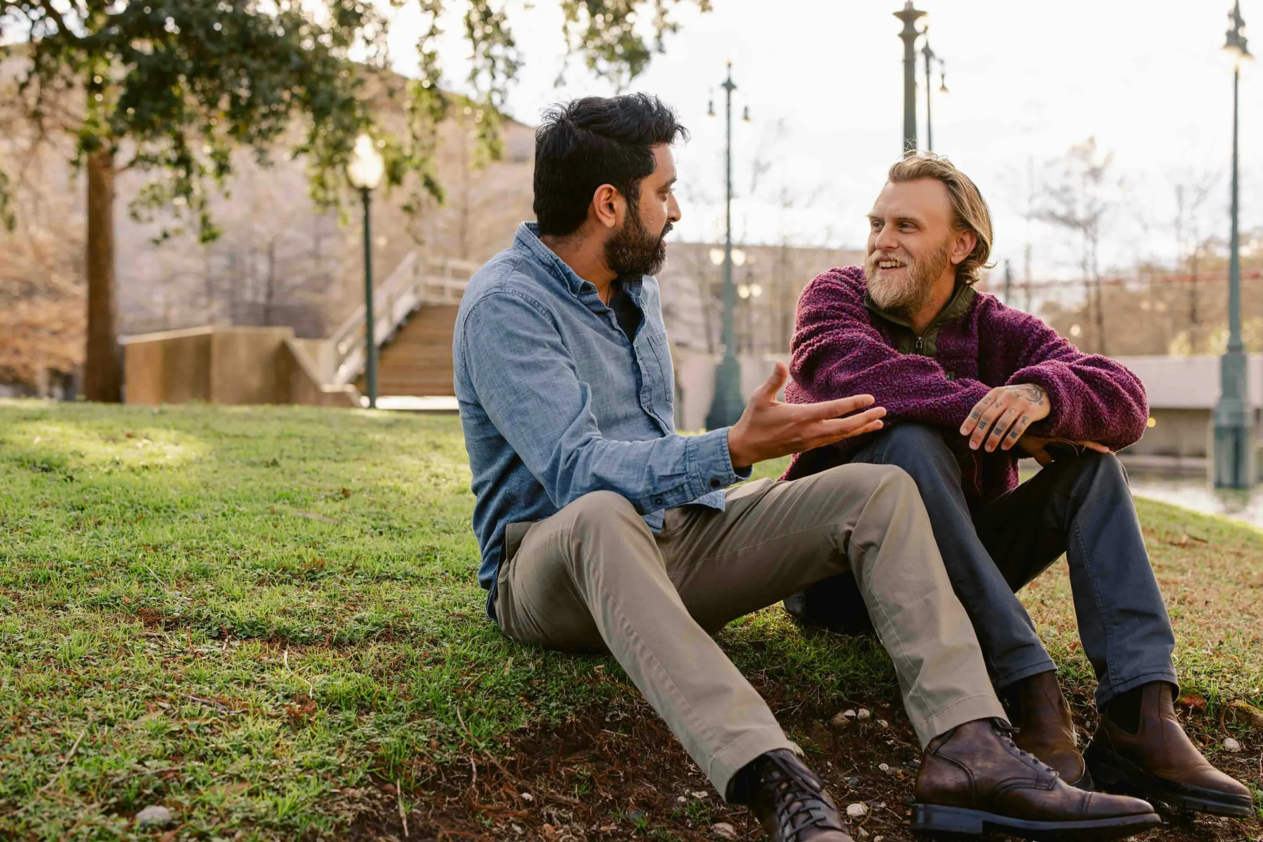 Two men from different cultural backgrounds engaged in animated, friendly conversation while sitting on grass in urban park setting, demonstrating successful cross-cultural communication