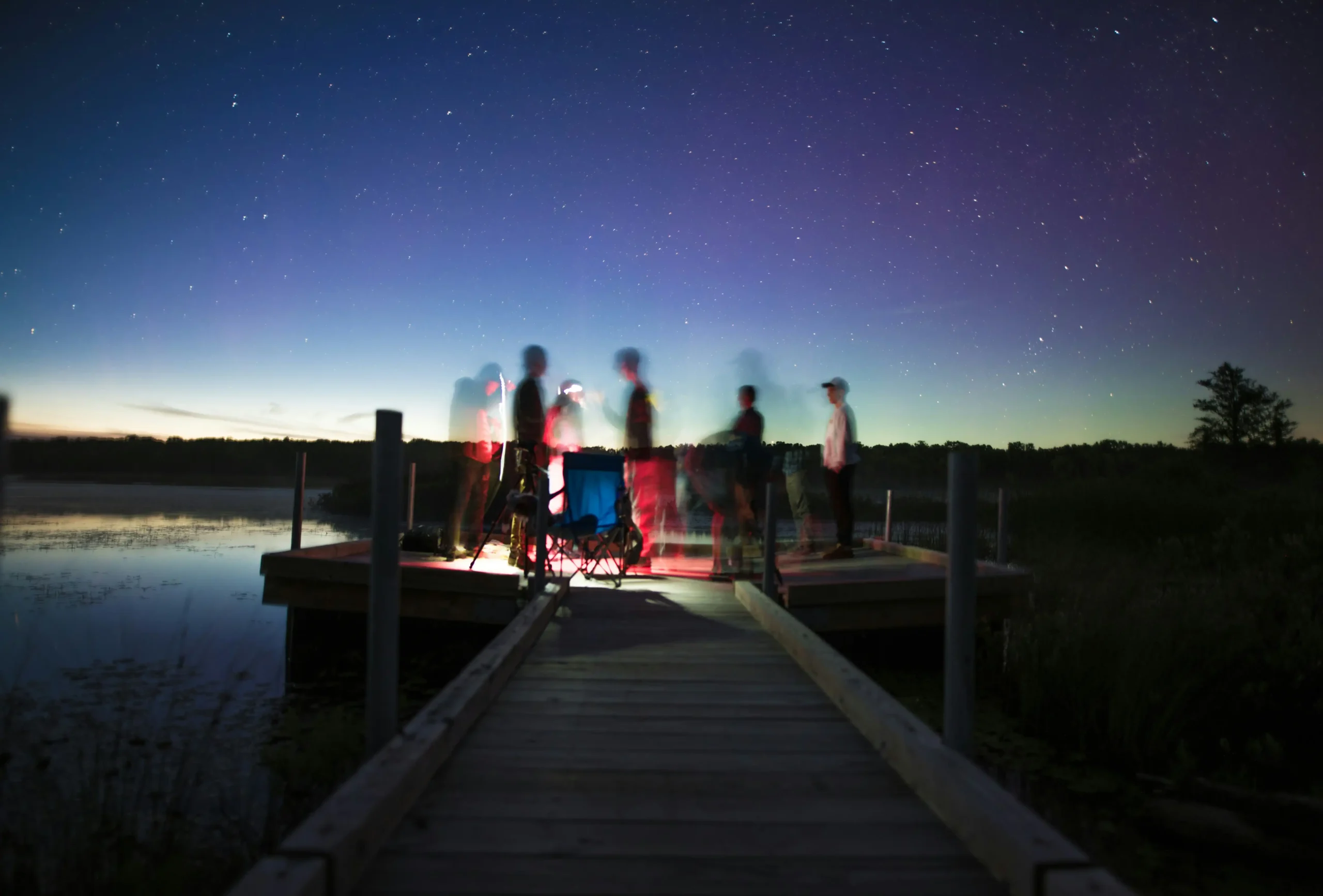 Group of diverse travelers gathered on wooden dock under starry twilight sky, sharing experiences around warm lighting by peaceful lake, representing community support for anxiety management
