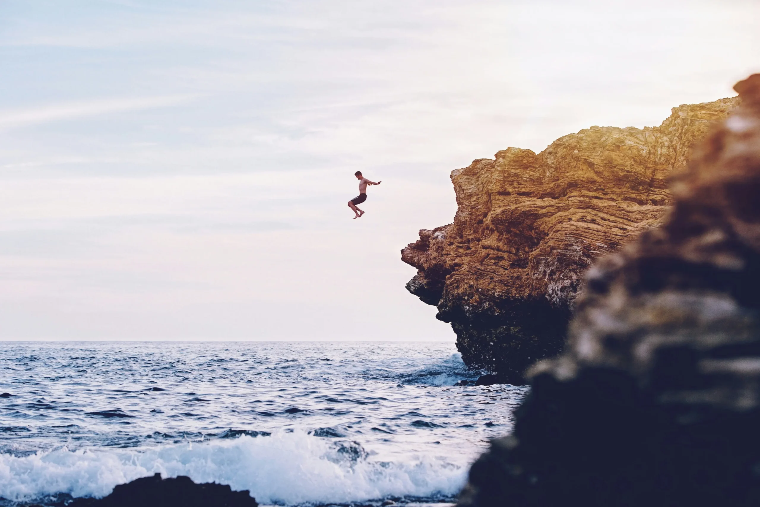 Person captured mid-jump from coastal cliff into crystal blue ocean water below rocky coastline
