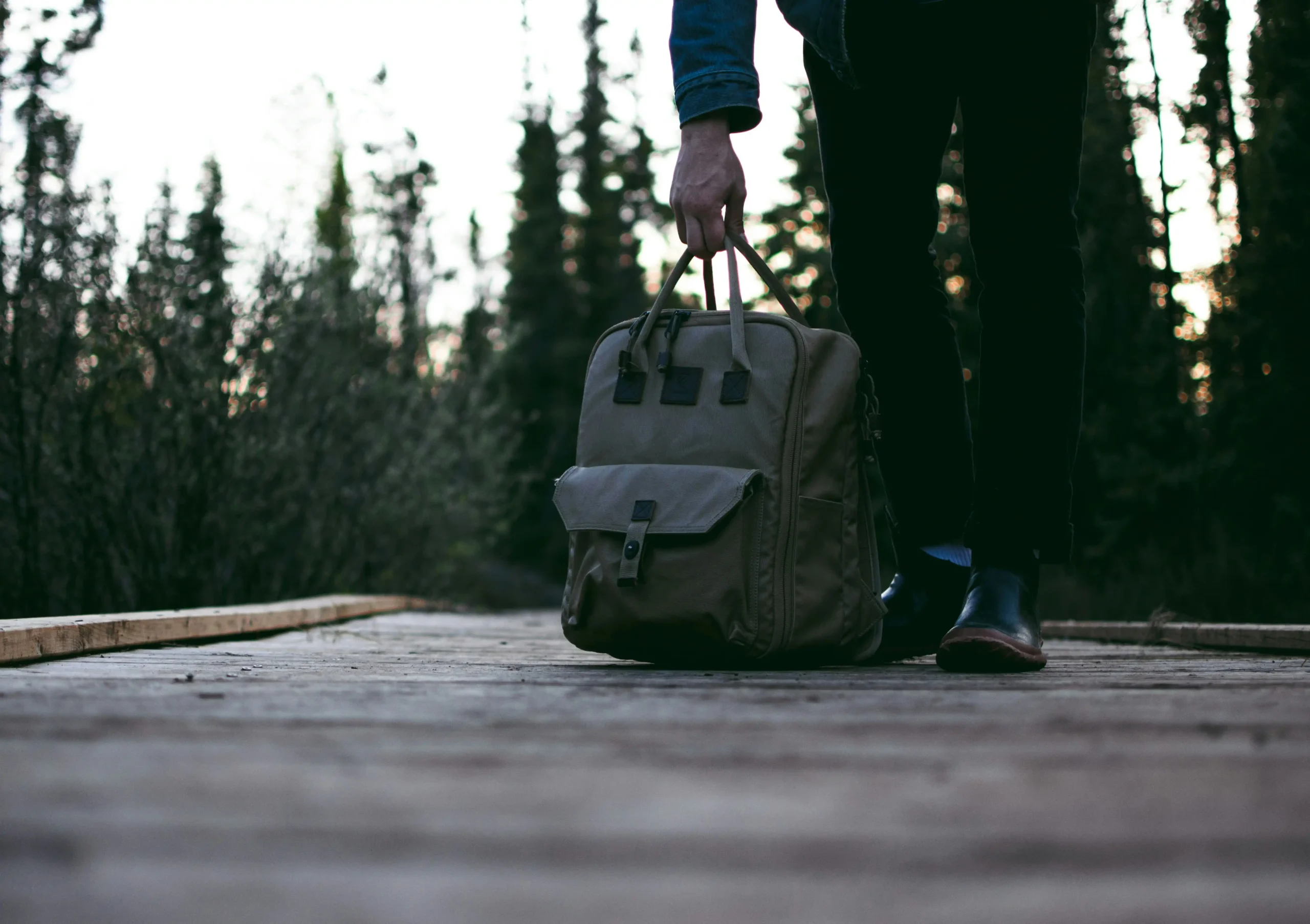 Person standing with canvas travel bag on wooden deck in forest setting with soft evening light filtering through trees