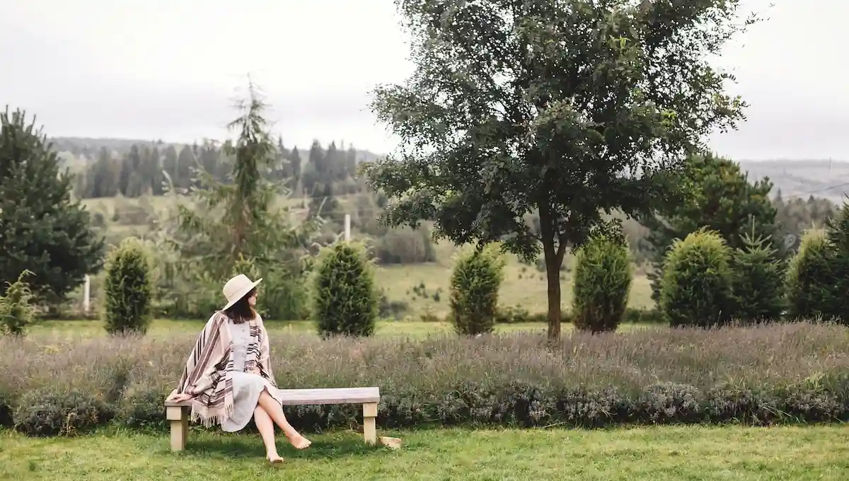  Woman in hat and shawl sitting peacefully on wooden bench in rural countryside overlooking rolling hills and farmland, embodying the unhurried mindfulness of slow travel