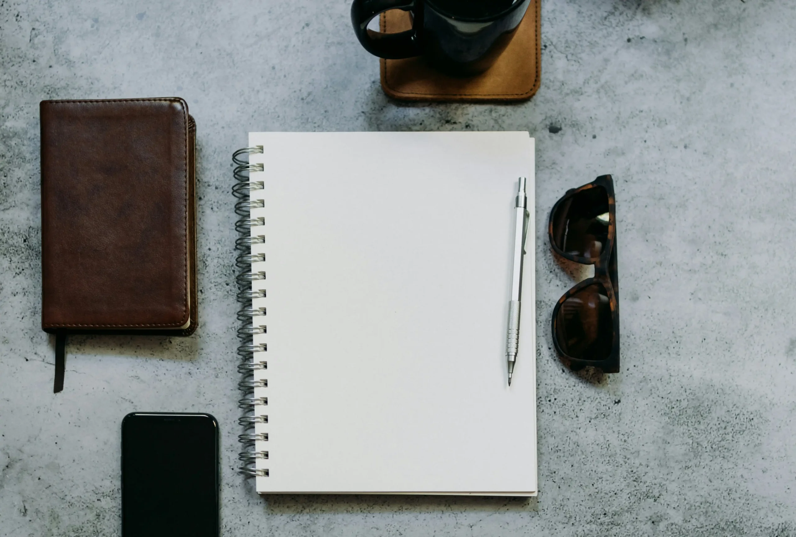 Clean workspace with blank notebook, pen, leather journal, sunglasses and coffee cup on concrete surface, representing mindful, intention-based travel planning