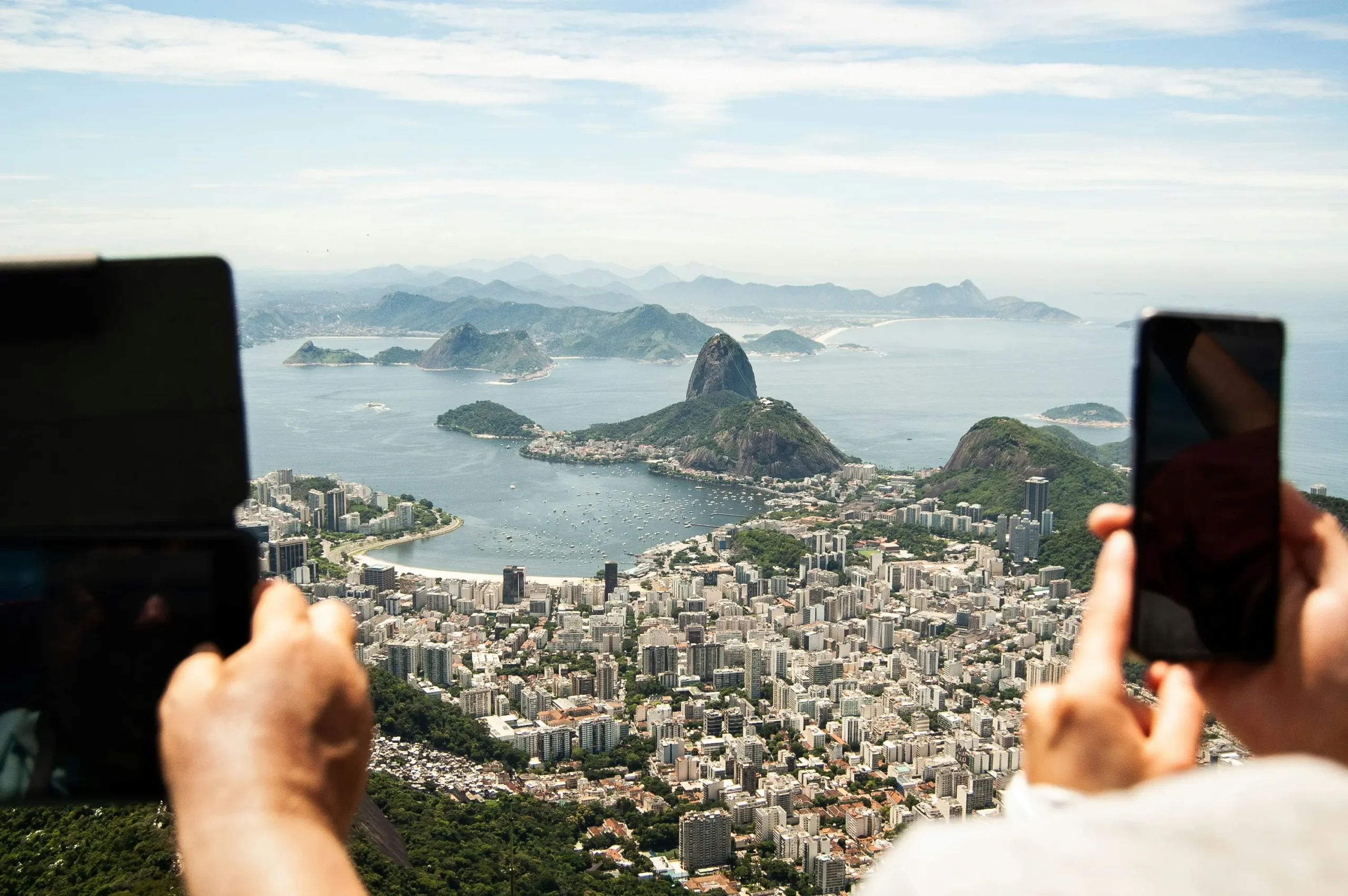 Multiple hands holding phones and tablets to photograph scenic Rio de Janeiro coastline with Sugarloaf Mountain and Guanabara Bay, showing mass documentation mentality