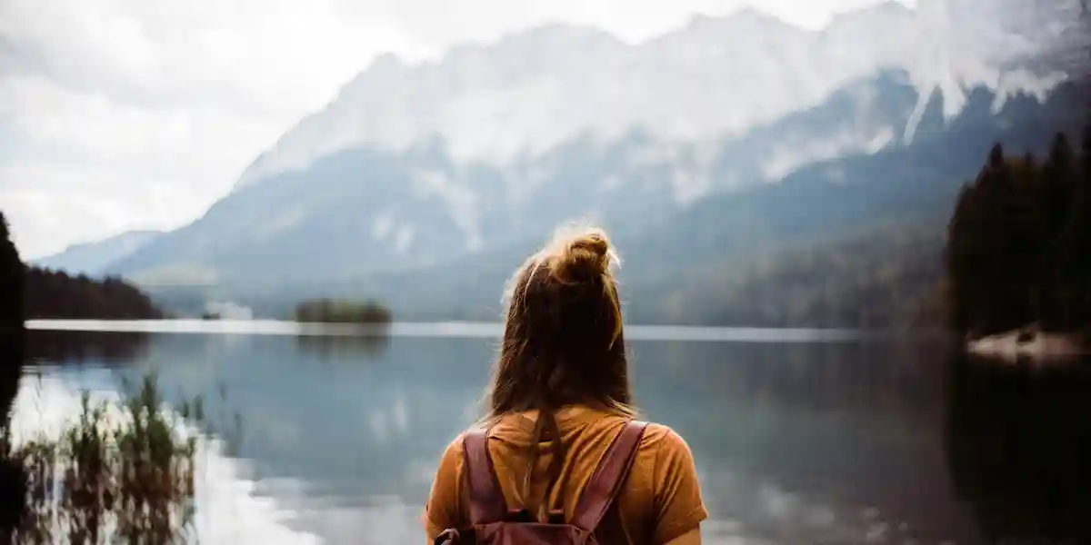 Woman with leather backpack standing peacefully at mountain lake shore, gazing at dramatic Alpine peaks reflected in calm water, representing the freedom and fulfillment of thoughtful budget travel