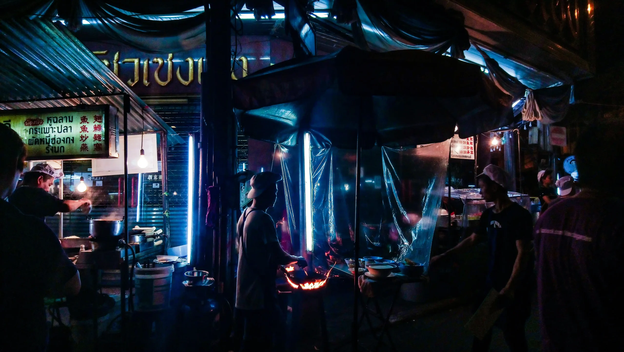 Atmospheric night street food market scene with vendors cooking under blue tarps and warm lighting, people gathering around food stalls