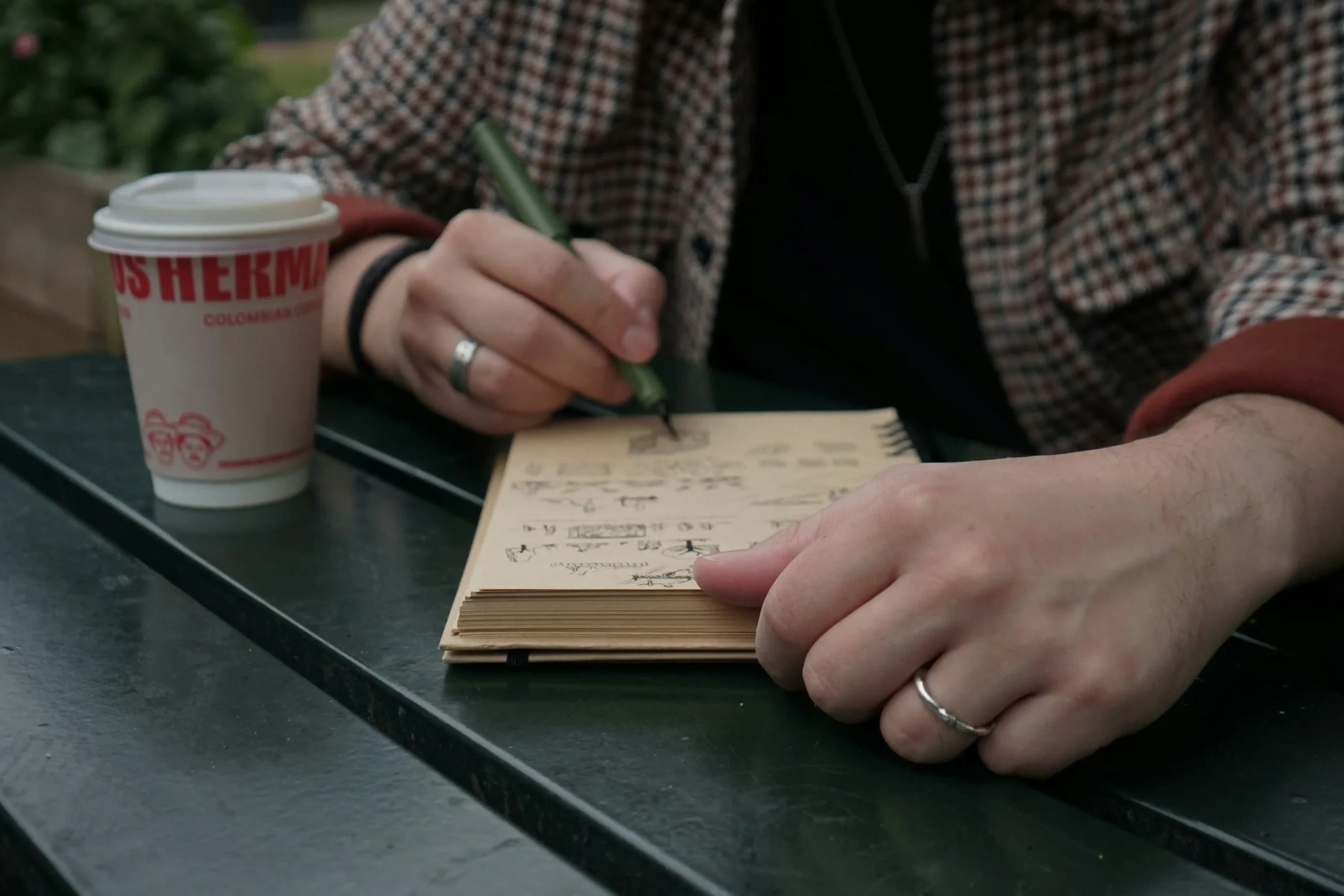  Person in plaid shirt writing thoughtfully in small notebook with coffee cup nearby at café table, representing the journaling and emotional preparation process for managing travel anxiety