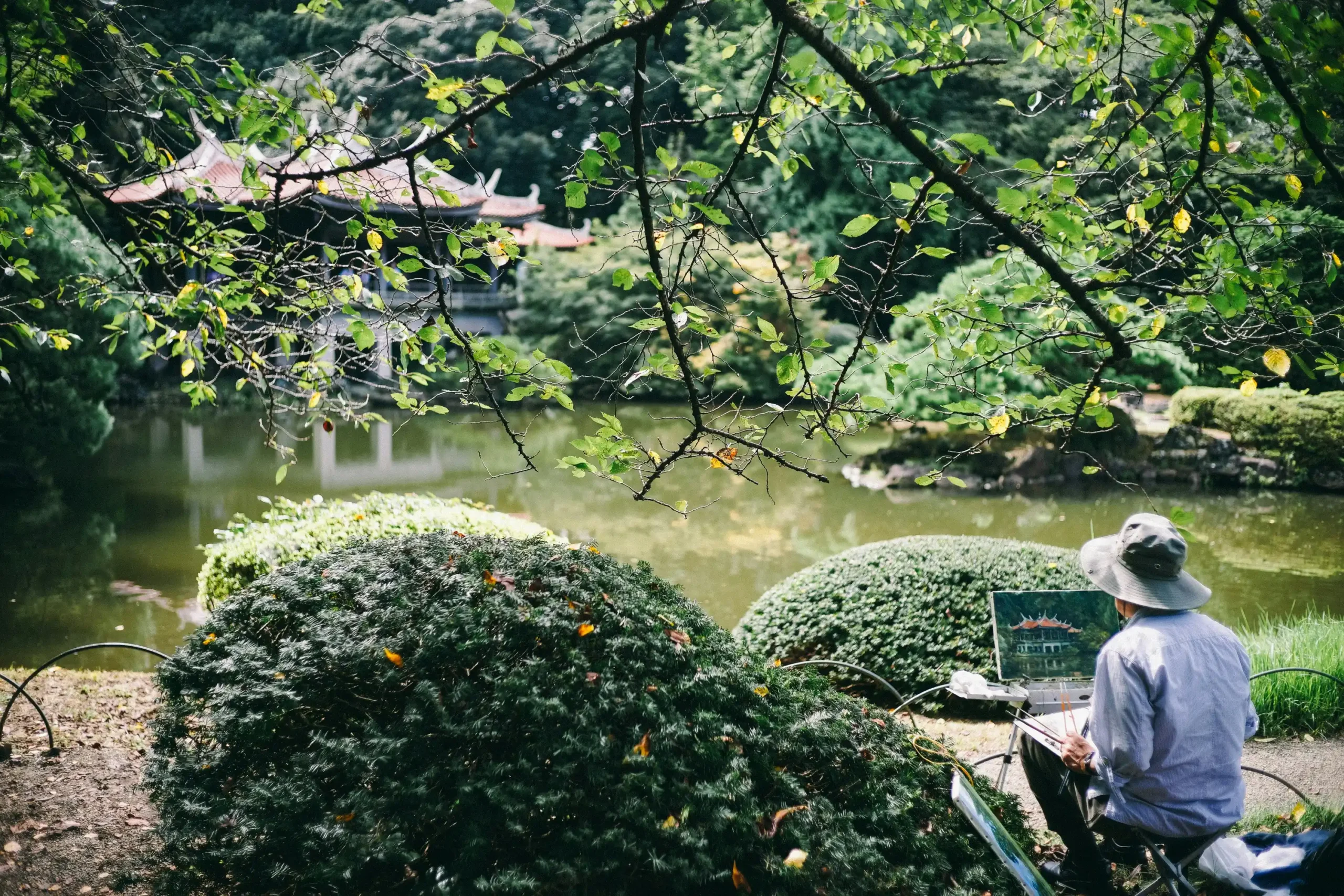 Person in hat sitting quietly by serene pond in Japanese garden, painting or sketching peacefully among carefully arranged rocks and overhanging branches
