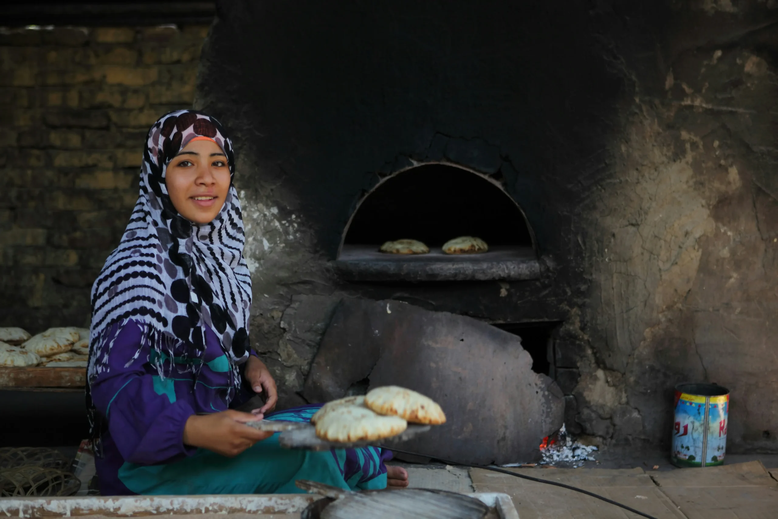 Woman in traditional headscarf baking fresh bread in ancient stone oven, representing the passing down of cultural traditions through hands-on participation