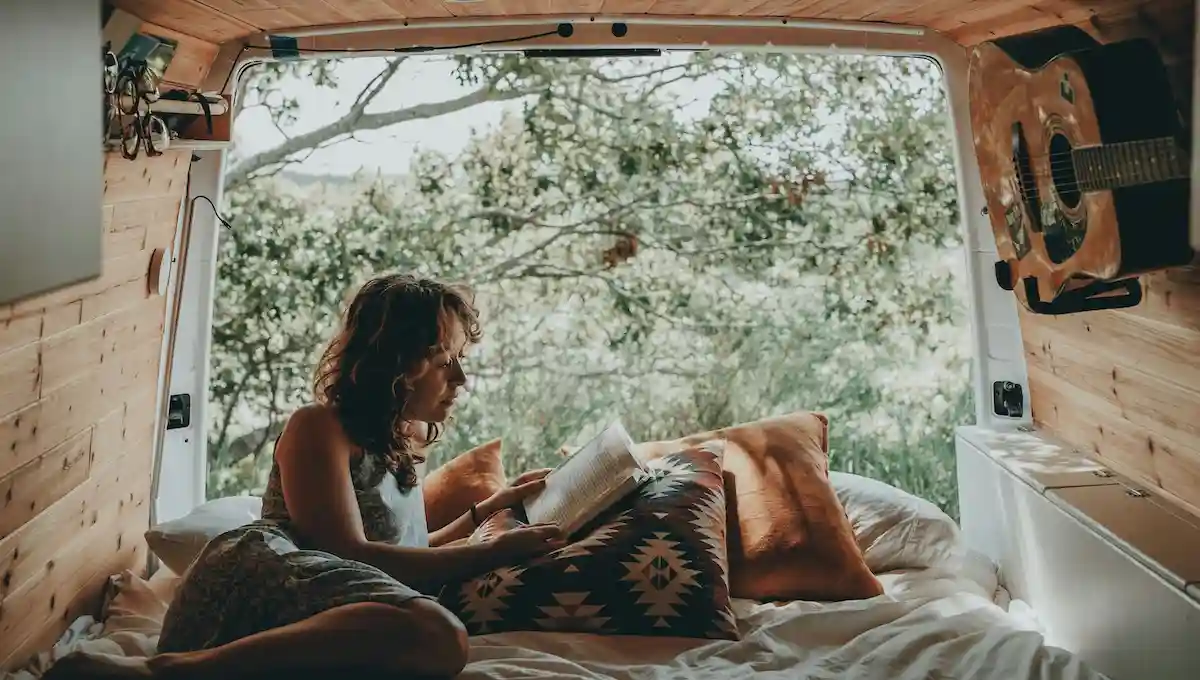  Woman reading thoughtfully in cozy van interior with natural light streaming through open doors, guitar and personal belongings visible, representing introspective travel planning focused on personal values rather than external research 