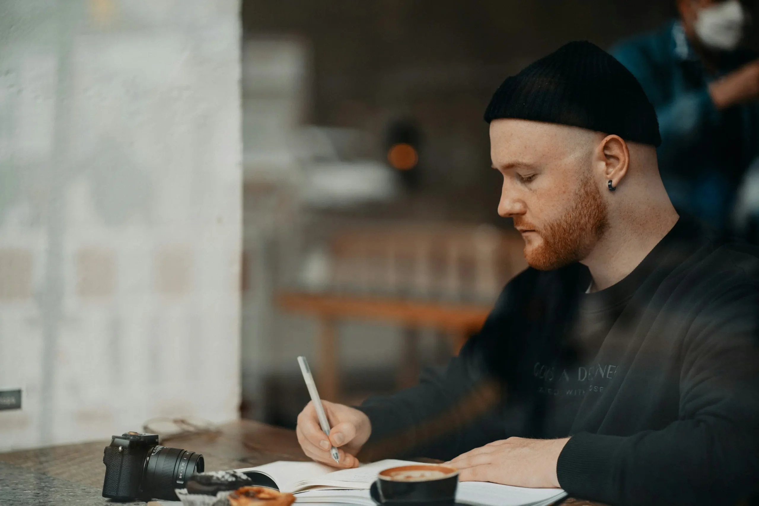 Man in beanie writing thoughtfully in notebook at coffee shop with camera and coffee nearby, focused on travel planning and reflection