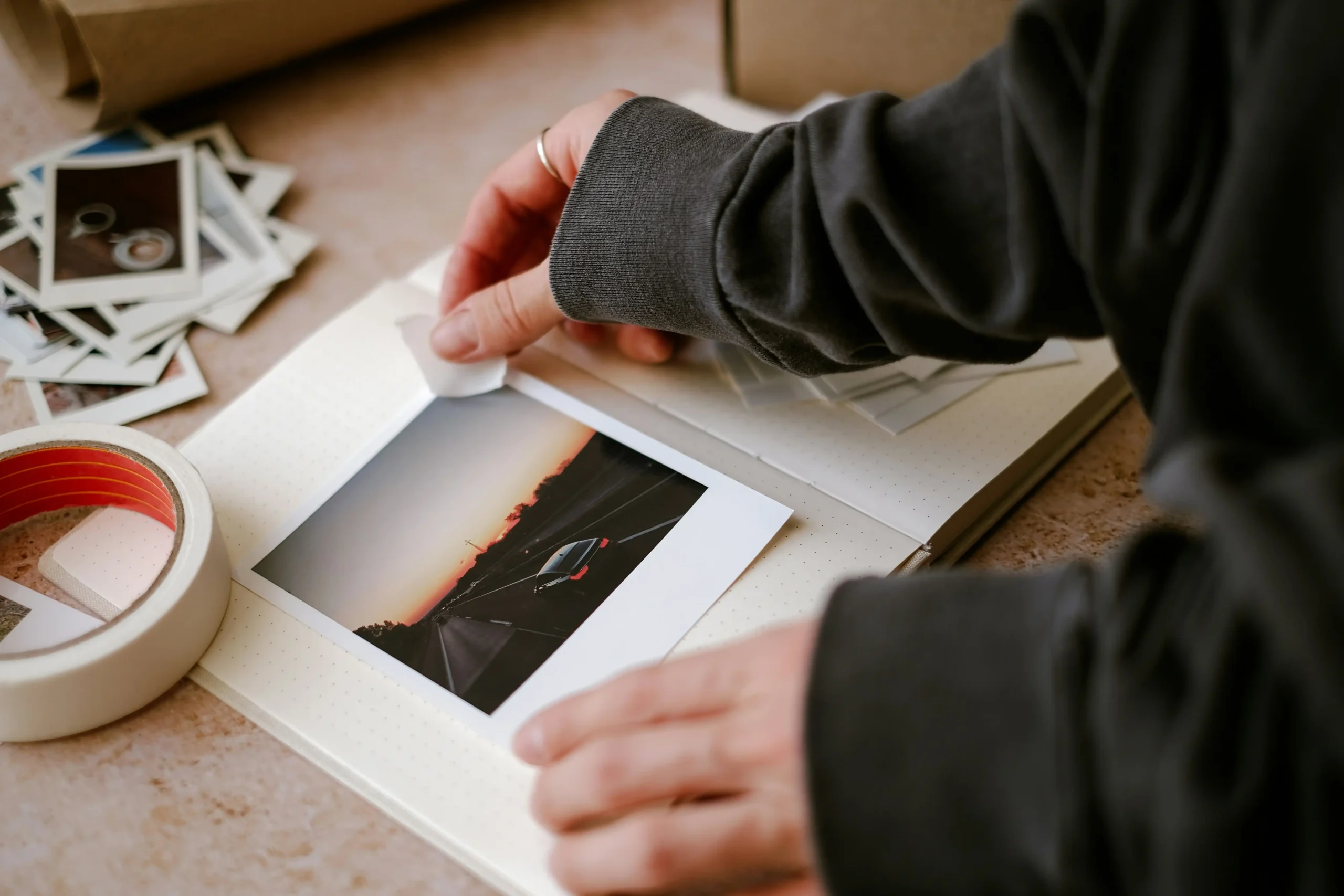  Person thoughtfully organizing travel photos in album at home with scattered instant photos nearby, representing post-travel reflection and integration of experiences