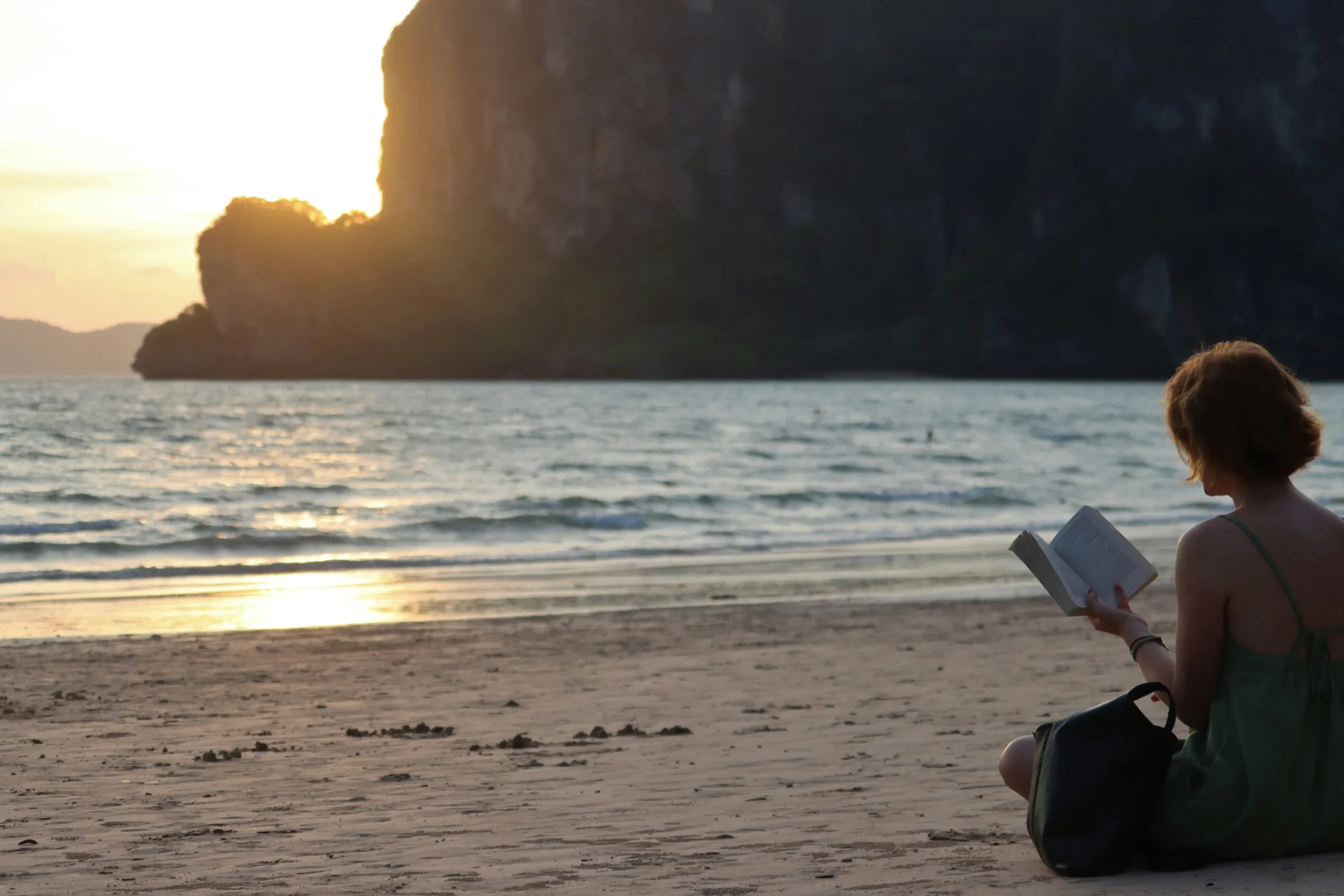 Woman reading book peacefully on beach at golden hour with dramatic coastal cliffs in background, embodying unhurried, contemplative travel