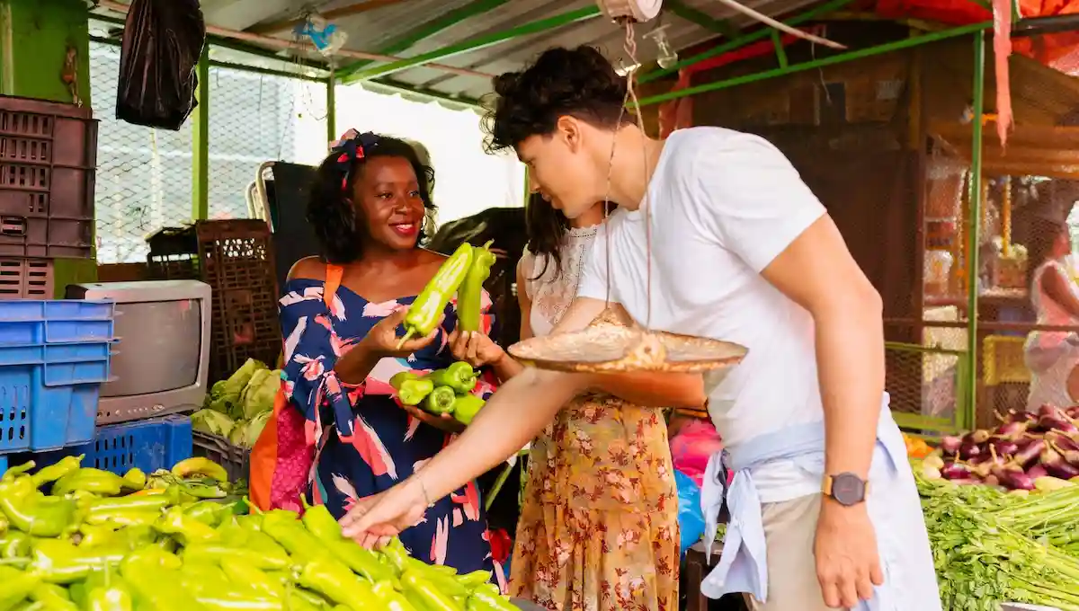 Young travelers shopping at vibrant local market, warmly interacting with friendly vendor surrounded by fresh green vegetables and tropical produce, showing genuine cultural exchange