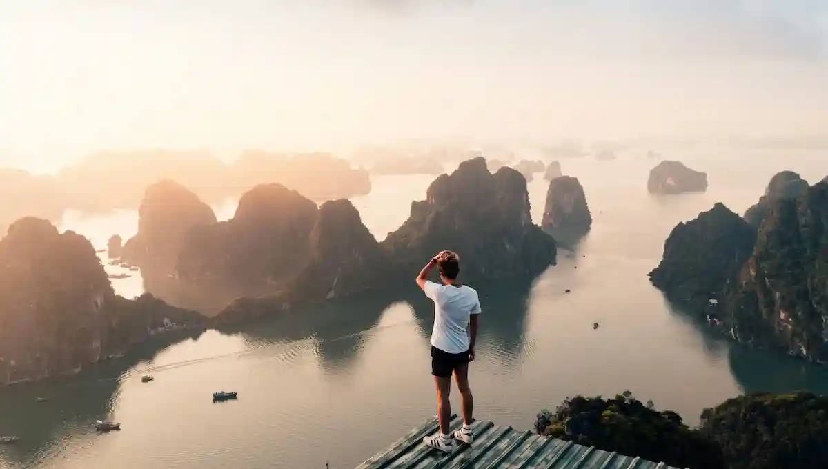 Person standing on wooden platform overlooking dramatic limestone karsts and emerald waters at golden hour, hand shading eyes while gazing into the distance, representing self-discovery and purposeful travel planning