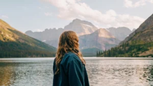 Person with flowing hair in denim jacket standing peacefully by pristine mountain lake, gazing at dramatic peaks reflected in calm water, representing serenity and overcoming travel anxiety