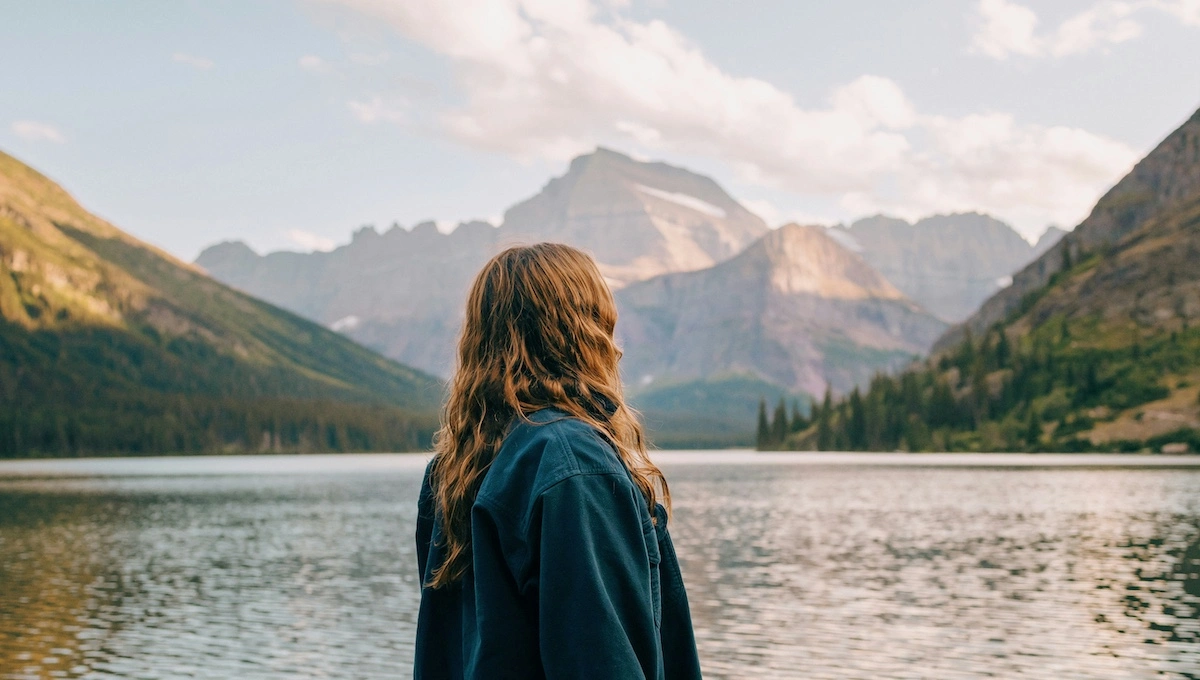 Person with flowing hair in denim jacket standing peacefully by pristine mountain lake, gazing at dramatic peaks reflected in calm water, representing serenity and overcoming travel anxiety