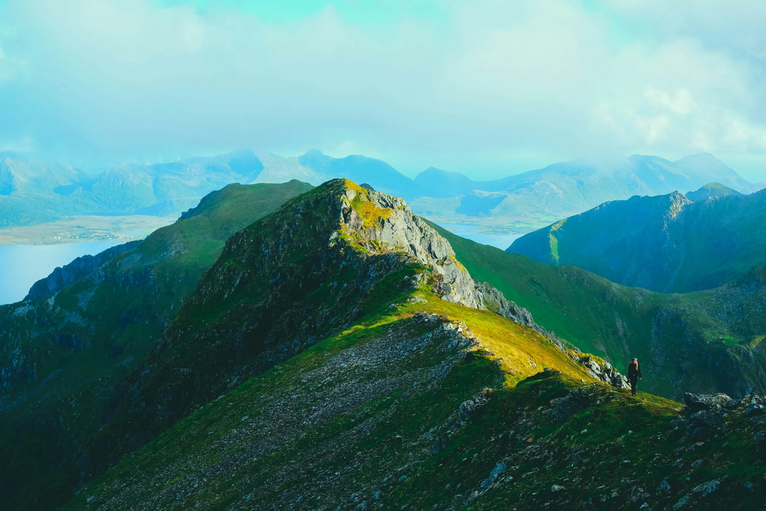 Lone figure in red jacket standing on dramatic mountain ridge with sweeping valley views and layered peaks in distance under vibrant sky