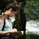 Young woman with backpack reading travel guide or map while standing in peaceful park setting with dappled sunlight filtering through trees