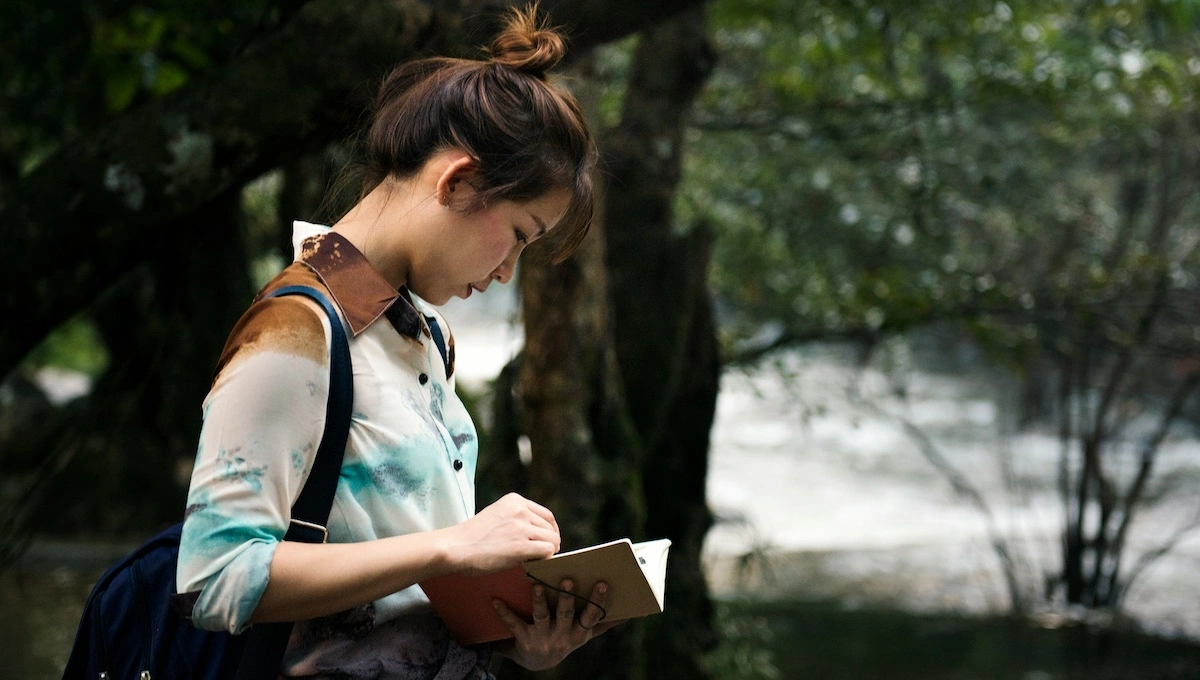 Young woman with backpack reading travel guide or map while standing in peaceful park setting with dappled sunlight filtering through trees