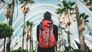 Traveler with red backpack walking through modern urban architecture with palm trees, representing someone ready for adventure through comprehensive preparation