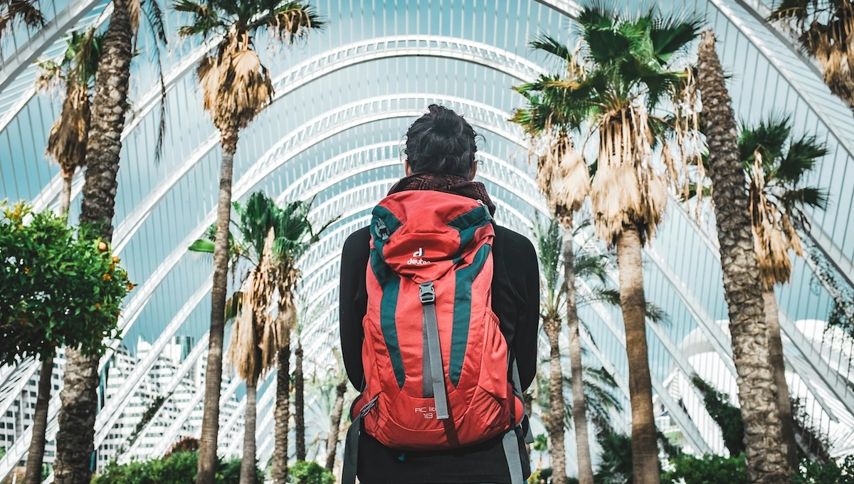 Traveler with red backpack walking through modern urban architecture with palm trees, representing someone ready for adventure through comprehensive preparation