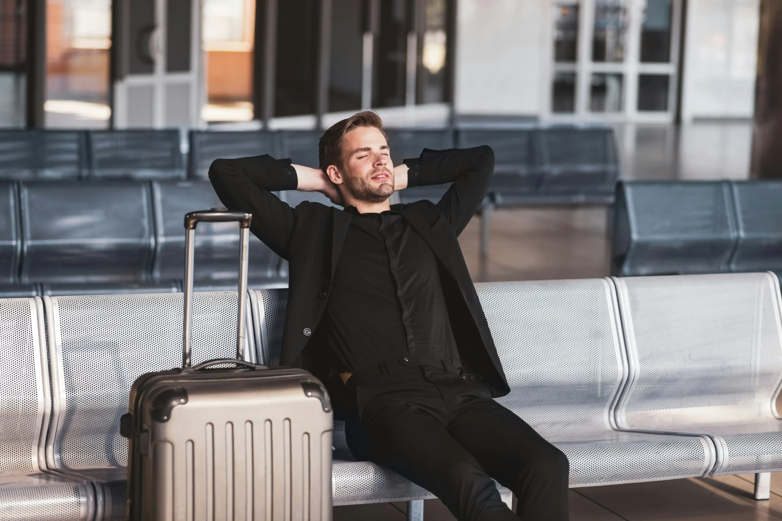 Man practicing relaxation technique with hands behind head and eyes closed while sitting peacefully in busy airport terminal with luggage nearby, demonstrating calm amid travel chaos