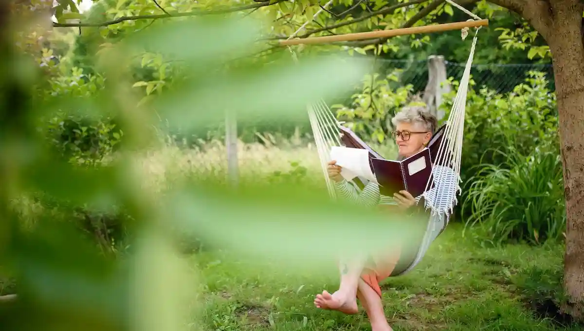 Woman with glasses reading contentedly in white hammock in lush garden setting, surrounded by green foliage and natural tranquility, embodying the rest and simplicity that come from knowing what you truly need 