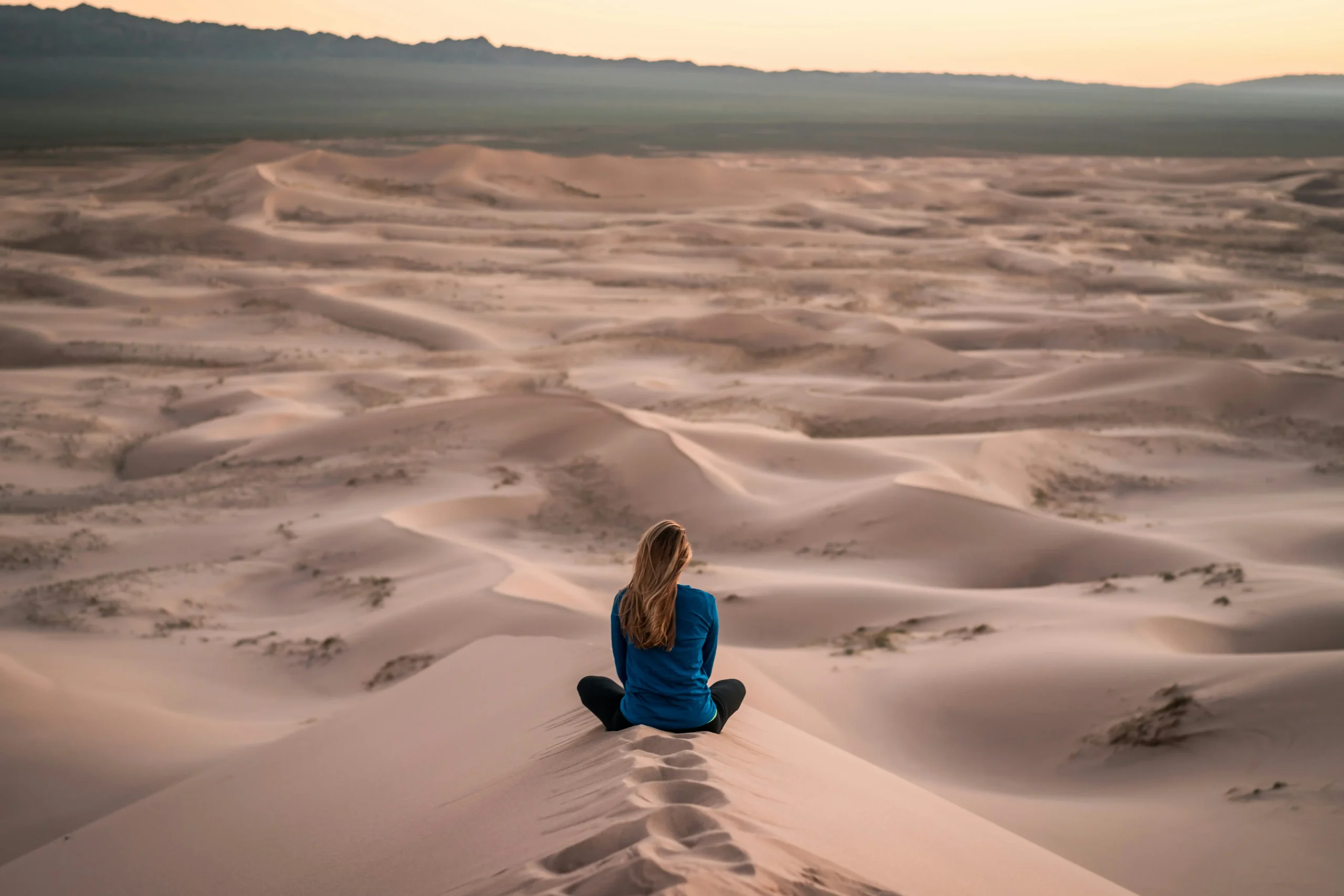 Lone female traveler sitting cross-legged on sand dune overlooking vast desert landscape at golden hour