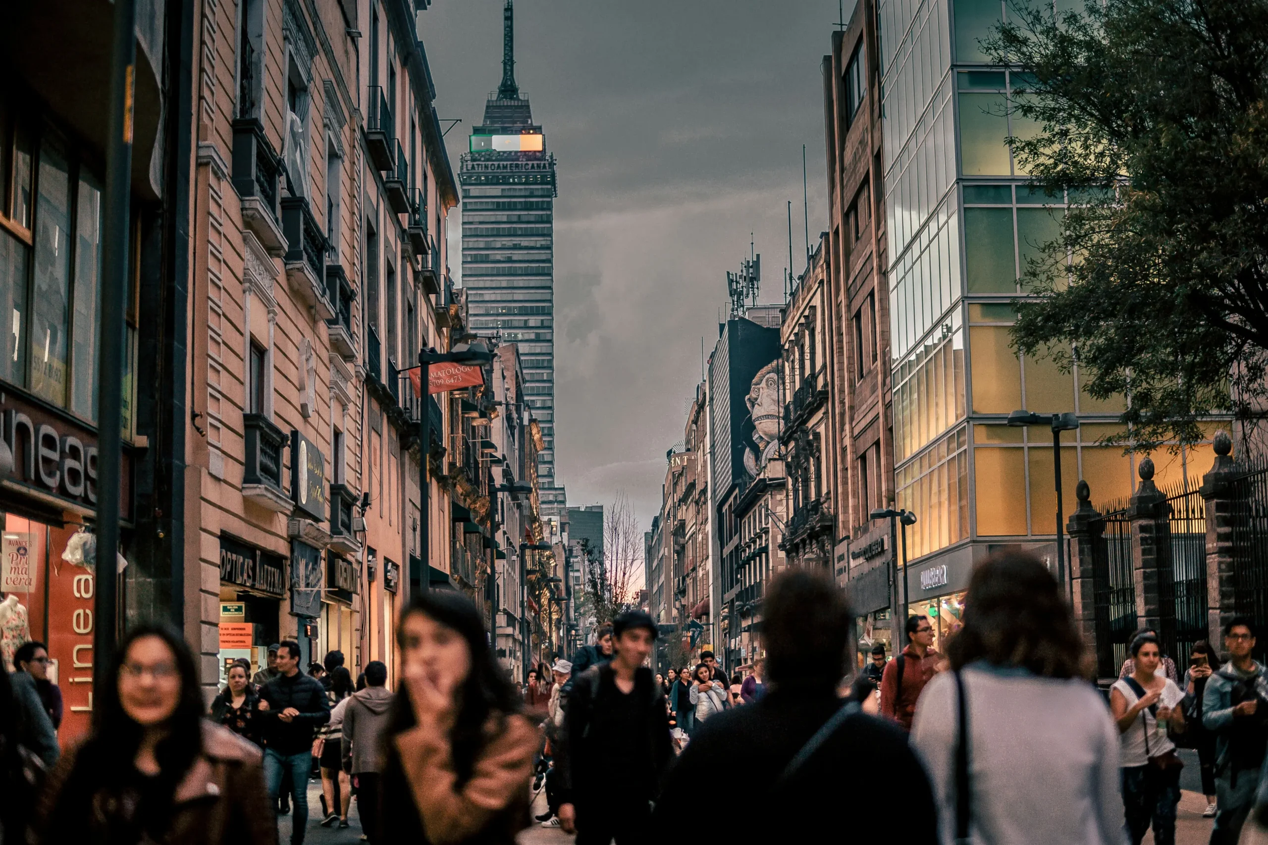 Crowded pedestrian street with motion-blurred people walking past historic buildings and shops during golden hour, with Torre Latinoamericana visible in background