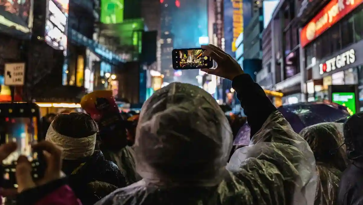  Person holding up phone to photograph busy neon-lit city street at night while surrounded by crowd of other tourists doing the same, representing social media-driven travel experiences