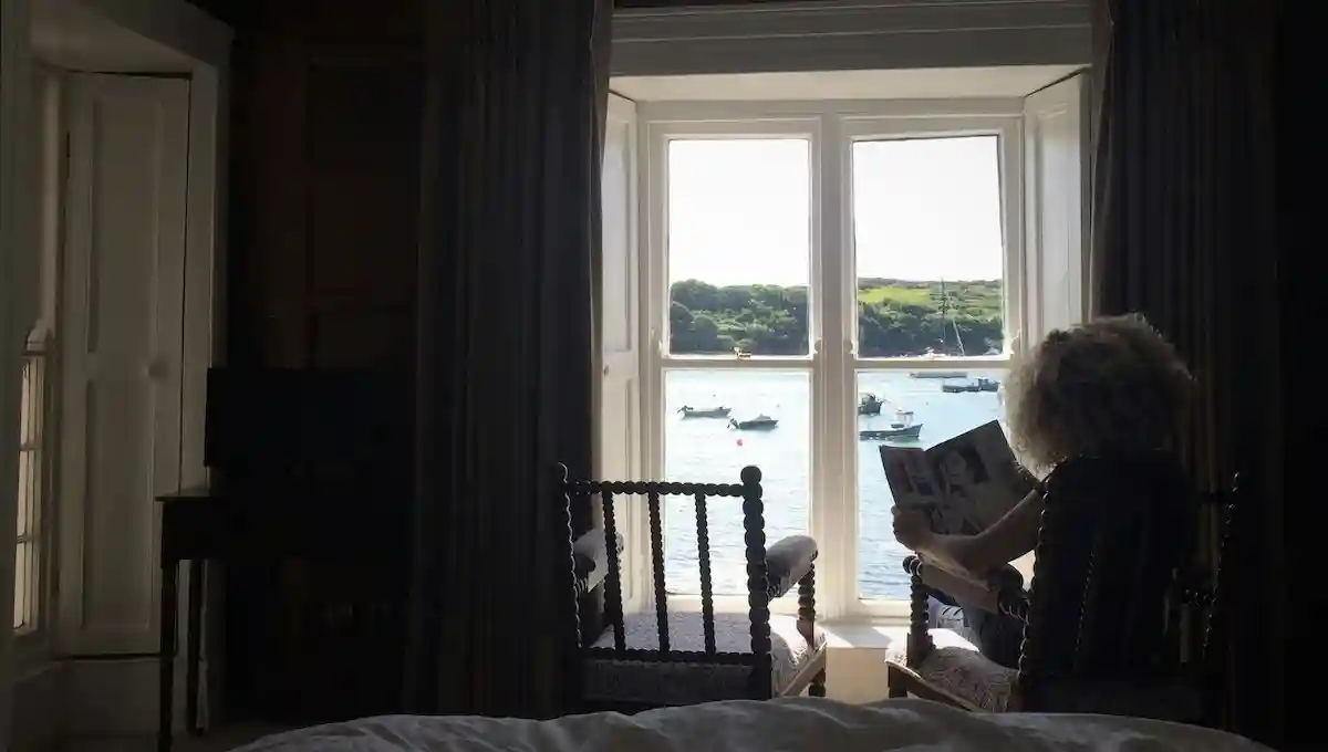 Person reading peacefully in comfortable guesthouse room with stunning harbor view through large windows, boats anchored in calm waters, representing the richness of longer, budget-conscious stays
