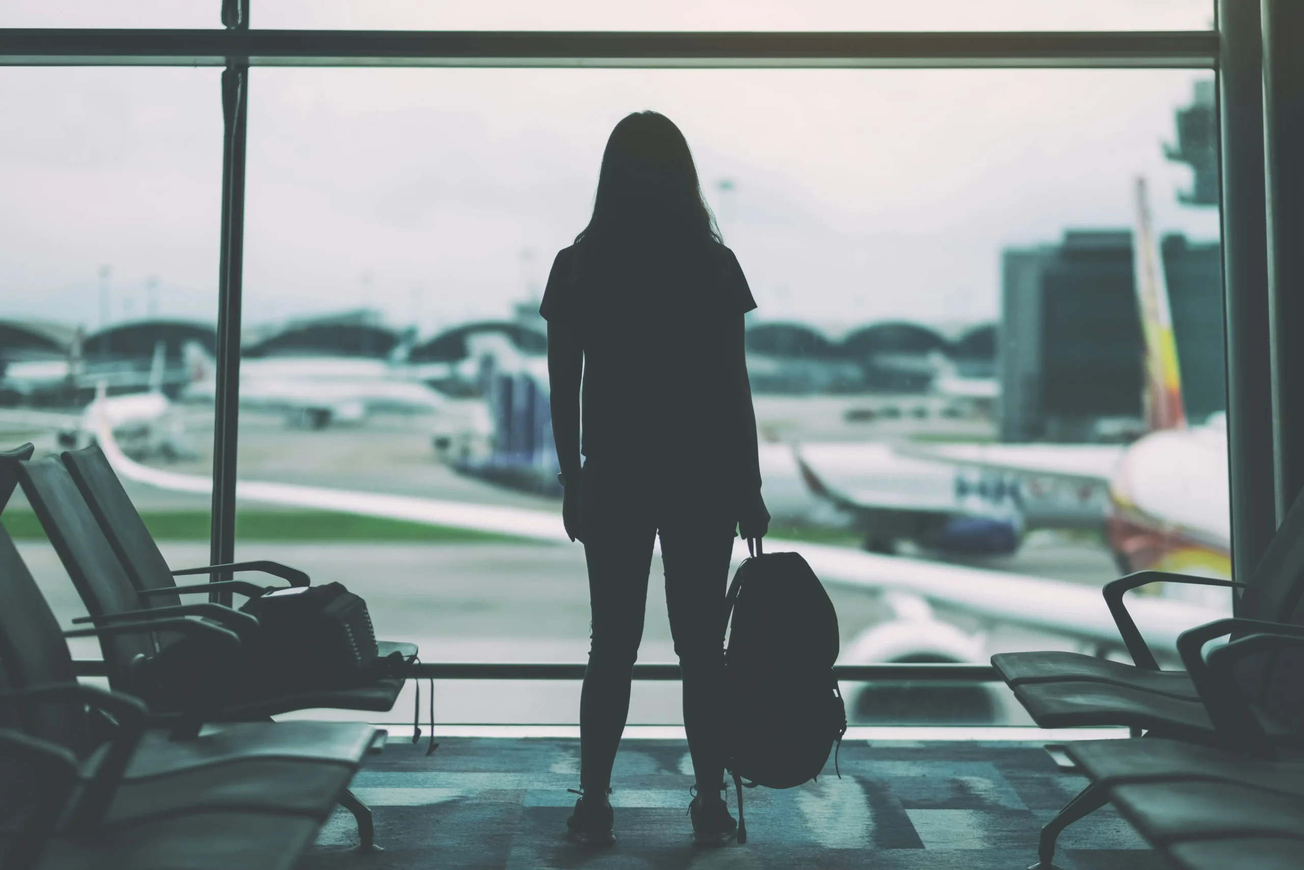  Silhouetted figure with luggage standing alone in airport terminal looking out at planes on tarmac, conveying contemplation and uncertainty